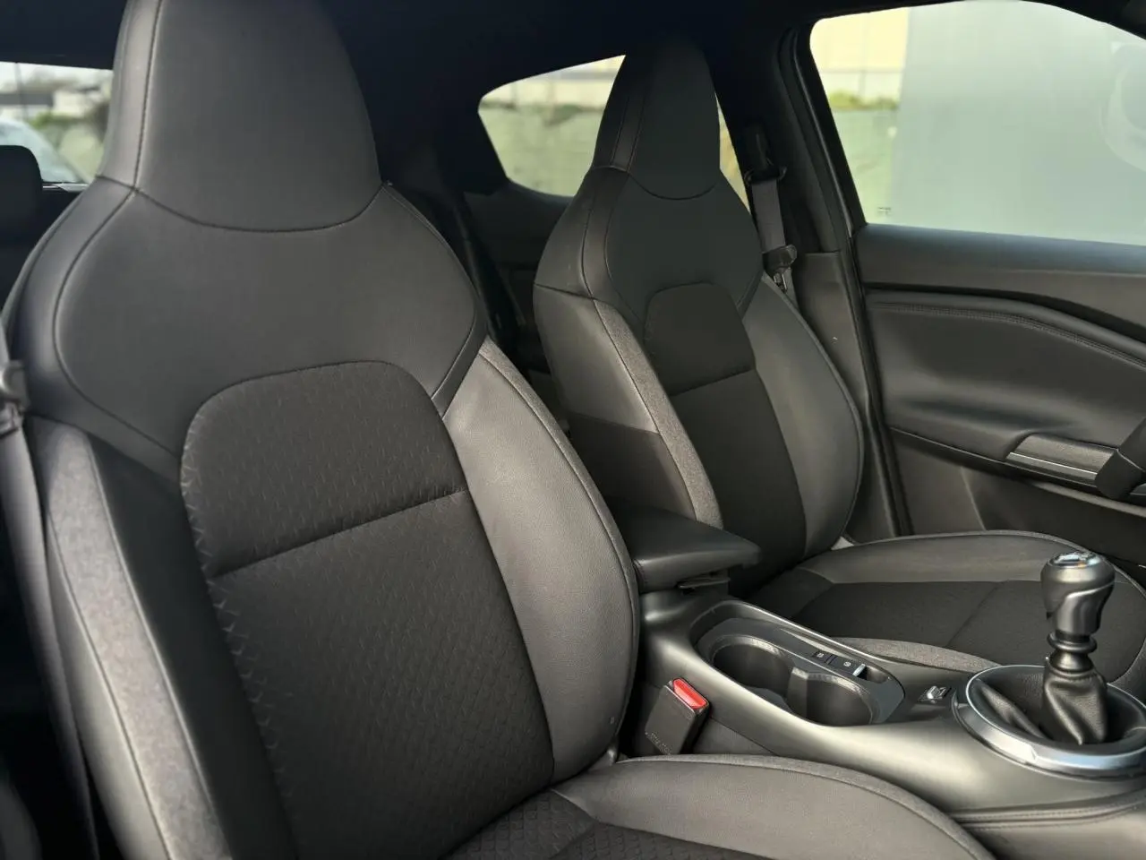 Vue intérieure côté avant droit du Nissan Juke 2025 avec sièges noirs en cuir et tissu, console centrale et levier de vitesses manuel.
