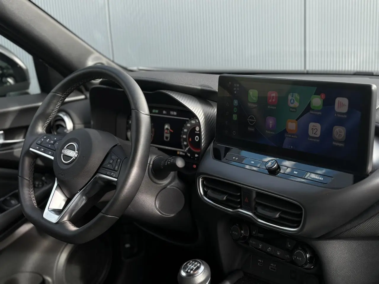 Vue intérieure côté conducteur du Nissan Juke 2025 gris foncé, volant cuir, écran tactile et levier de vitesse manuel.