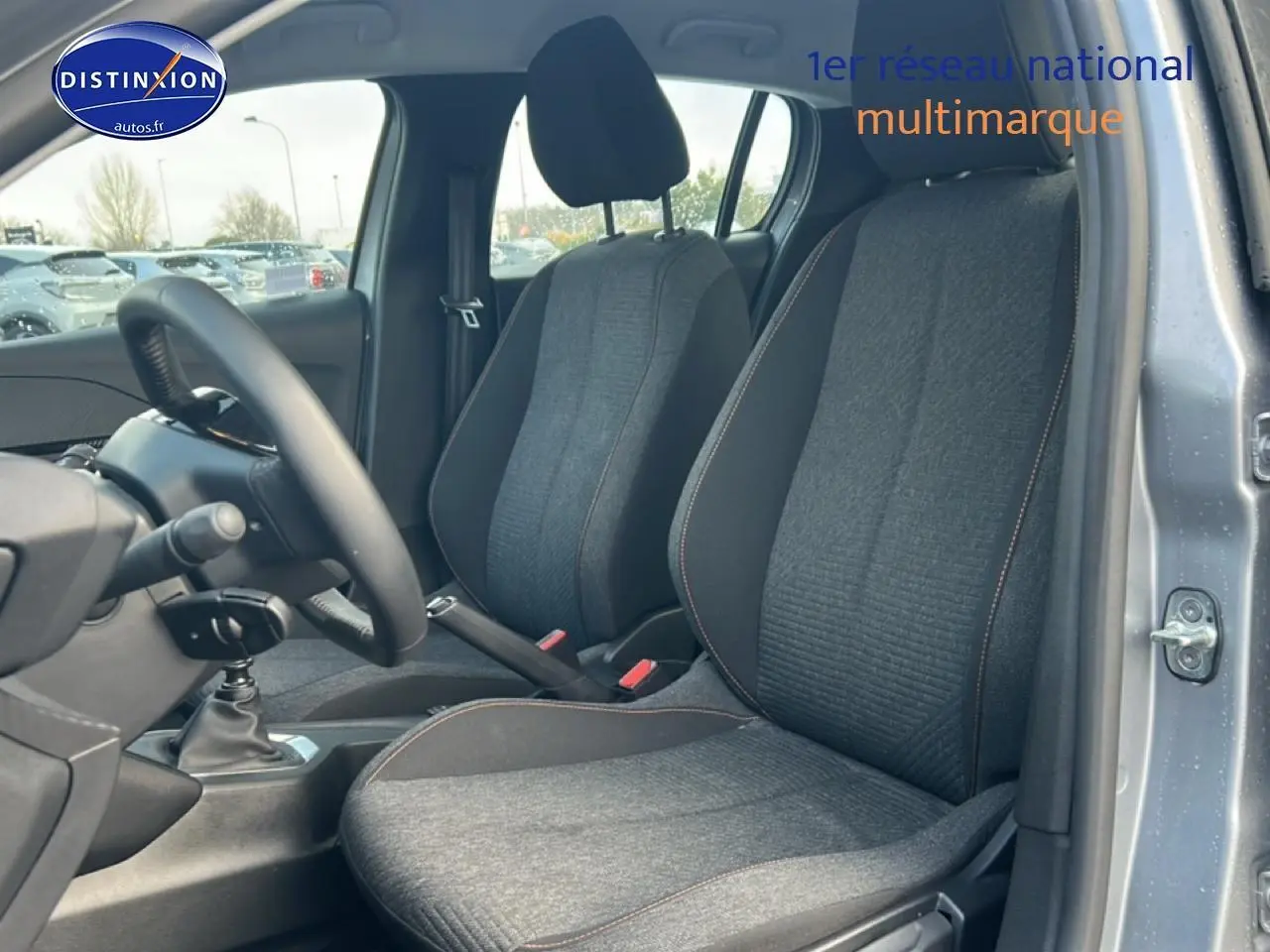 Vue intérieure côté avant droit de la Peugeot 208 1.2 100 Style 2025, sièges tissu gris avec surpiqûres orange.