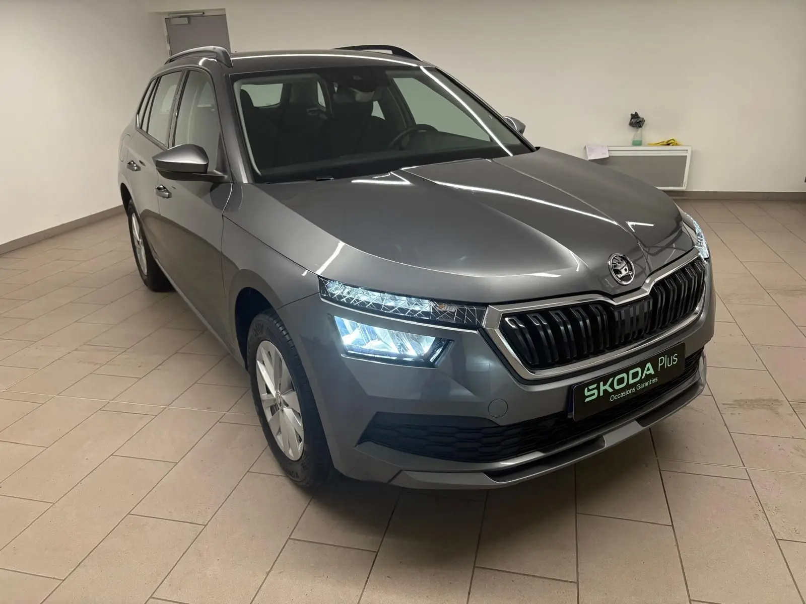 Vue 3/4 avant droite d'un Skoda Kamiq gris graphite avec phares allumés dans un intérieur neutre.
