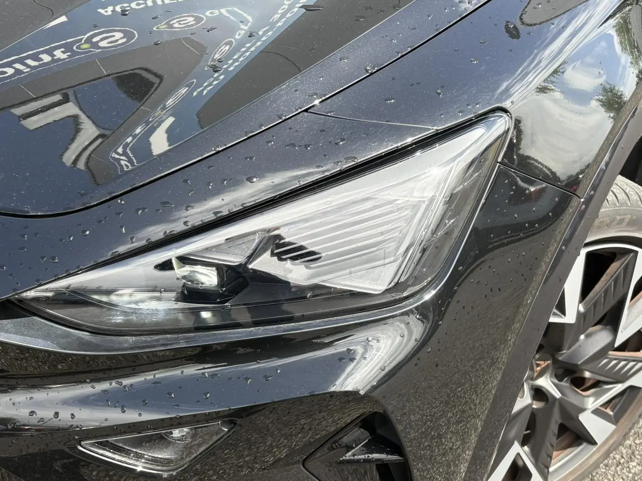 Gros plan sur le phare avant droit et la jante alliage 19'' Sandstorm du CUPRA Formentor Noir Minuit, avec gouttes d'eau.