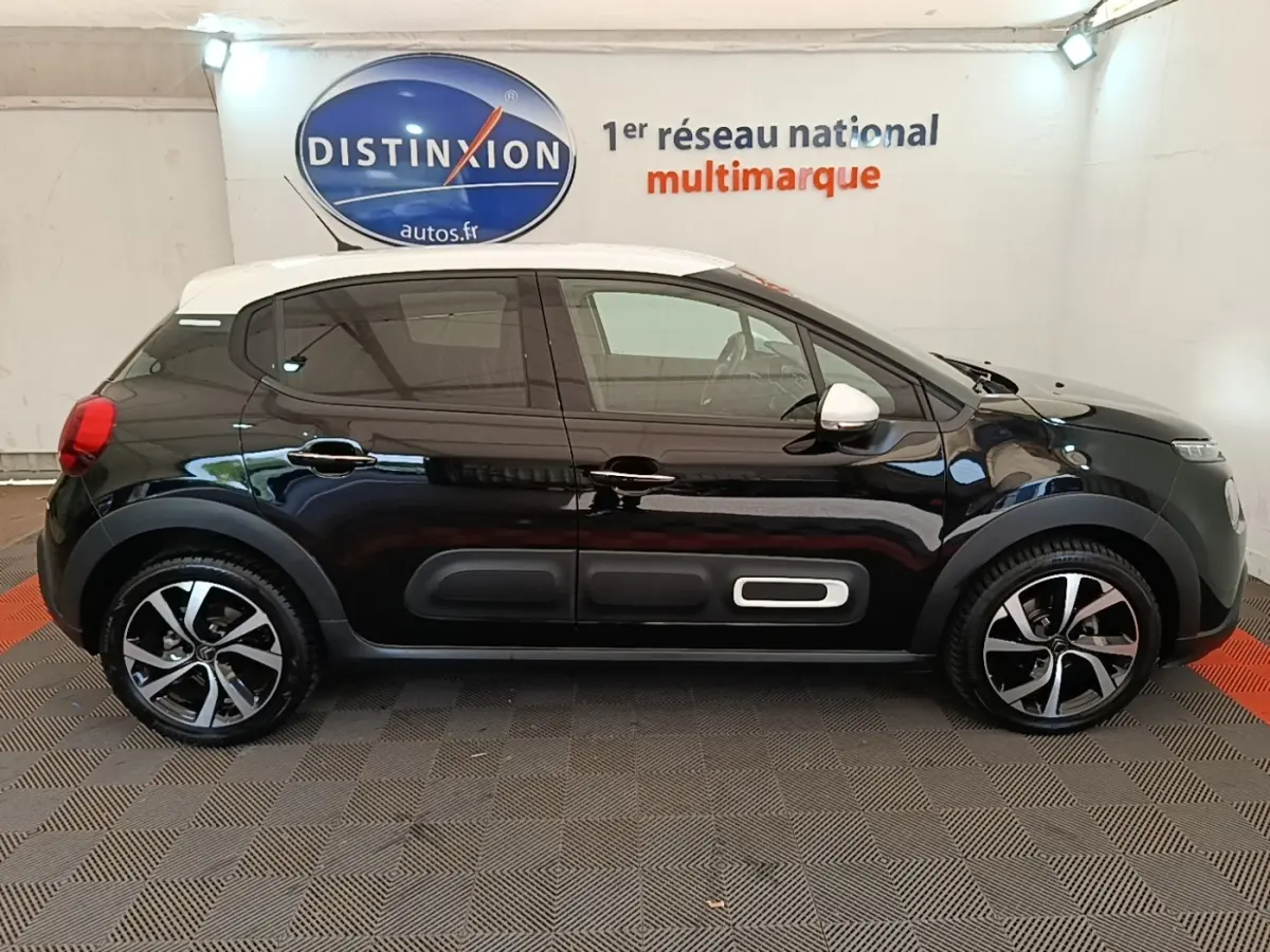 Vue de profil droit d'une Citroën C3 noire avec toit blanc et jantes alliage bicolores dans un showroom.