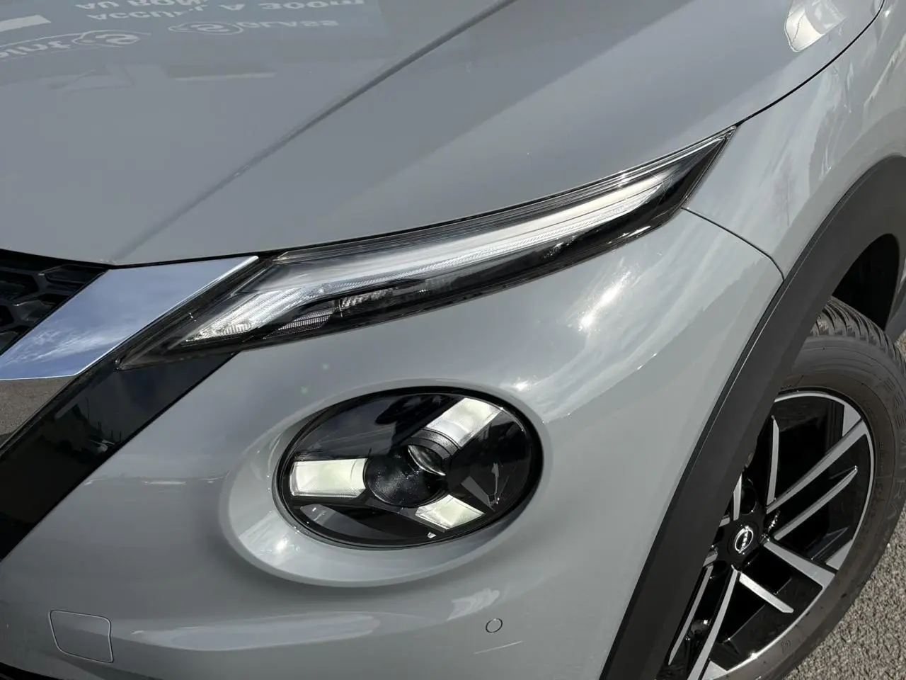 Gros plan sur le phare avant droit et la roue d’une Nissan Juke gris Ceramic, mettant en valeur les détails lumineux et la jante noire.