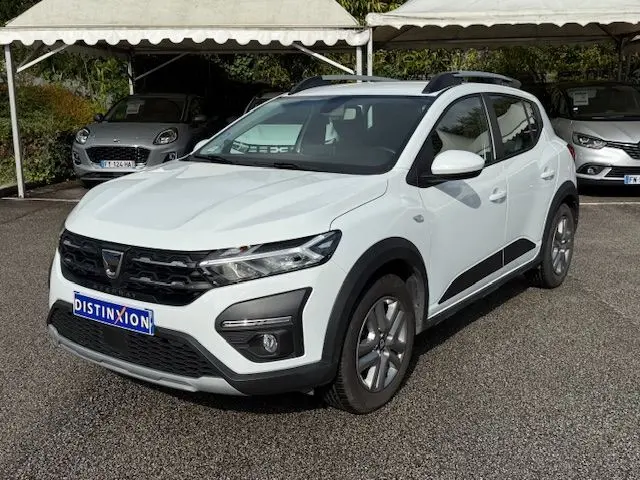 Vue 3/4 avant d'une Dacia Sandero Stepway blanche 2021 avec barres de toit et protections noires sur les passages de roue.