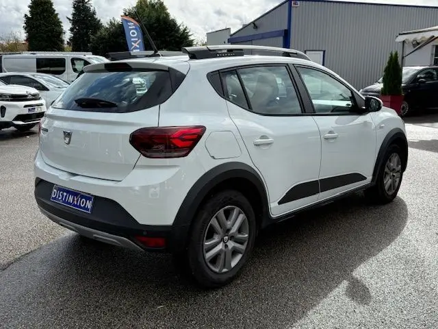 Vue 3/4 arrière droite d'une Dacia Sandero Stepway blanche 2021 avec barres de toit et protections latérales noires.