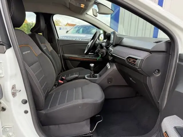 Vue intérieure côté conducteur d'une Dacia Sandero Stepway blanche 2021, sièges noirs avec surpiqûres orange et tableau de bord moderne.