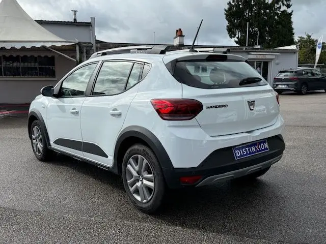 Vue 3/4 arrière droite d'une Dacia Sandero Stepway blanche 2021 avec barres de toit et protections noires sur les bas de caisse.