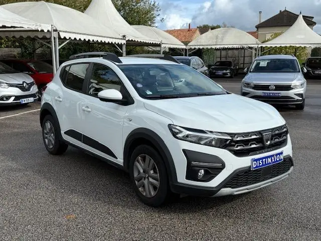 Dacia Sandero Stepway blanc vue 3/4 avant droit, avec barres de toit et protections noires sur les bas de caisse.