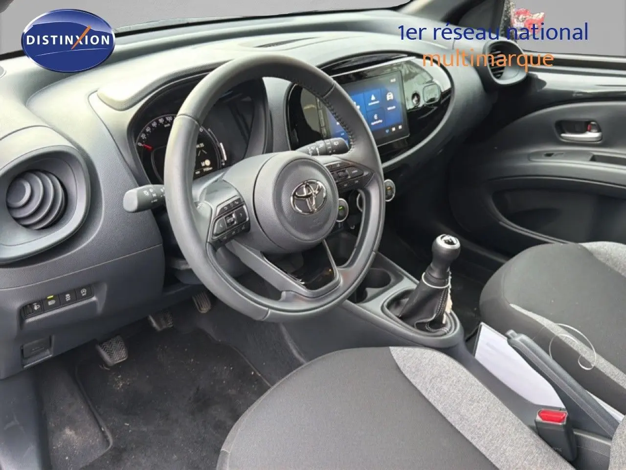 Vue intérieure de face du poste de conduite noir de la Toyota Aygo X 2025 avec volant multifonction et boîte manuelle.