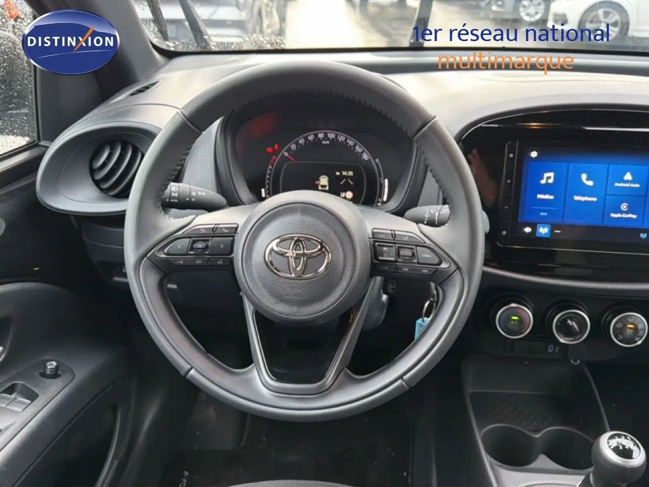 Vue intérieure centrée sur le volant et le tableau de bord noir de la Toyota Aygo X 2025 avec écran tactile et commandes au volant.