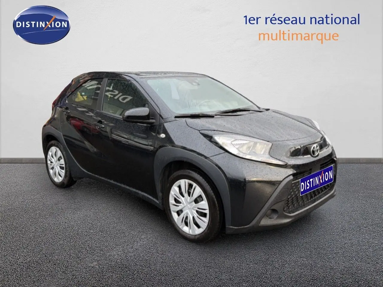 Toyota Aygo X noir vue 3/4 avant droit avec calandre distinctive et jantes argentées sur sol gris.