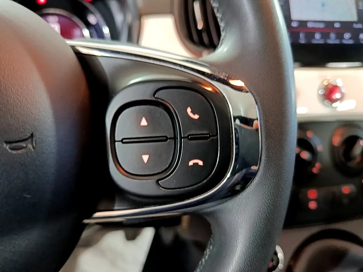 Gros plan sur les commandes du volant cuir noir de la Fiat 500 Hybrid 2021, avec tableau de bord flou en arrière-plan.