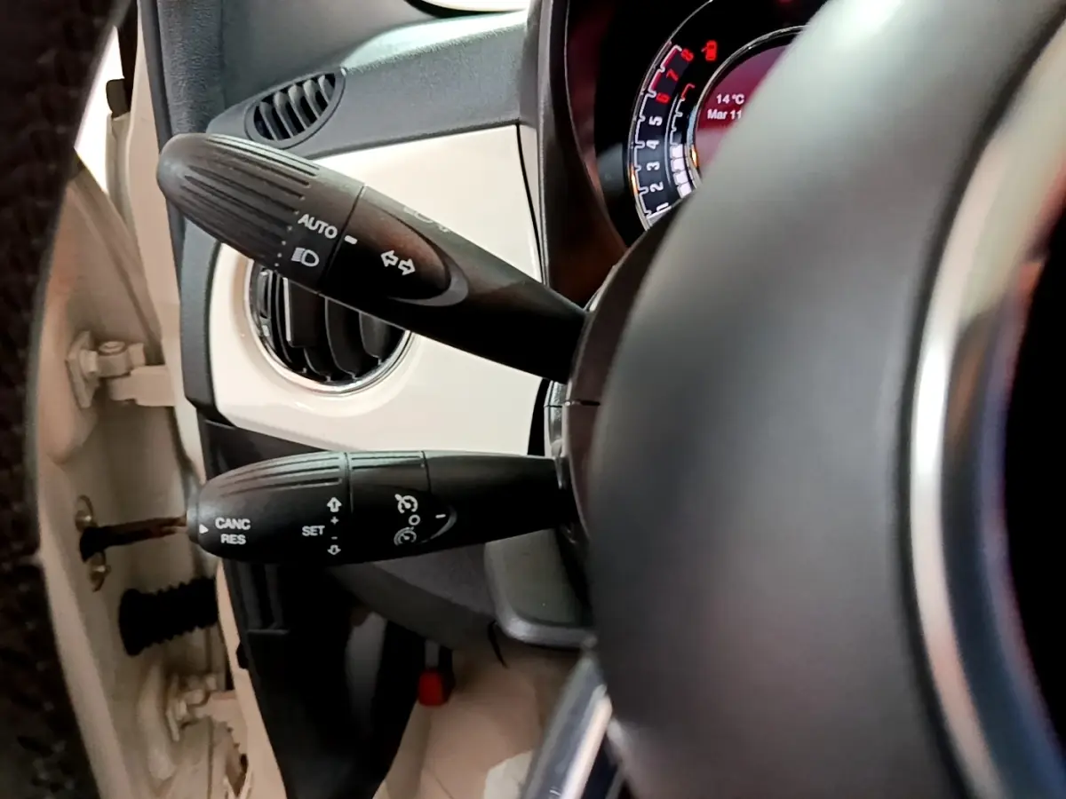 Gros plan sur la colonne de direction et les commandes au volant d’une Fiat 500 blanche, tableau de bord partiellement visible.