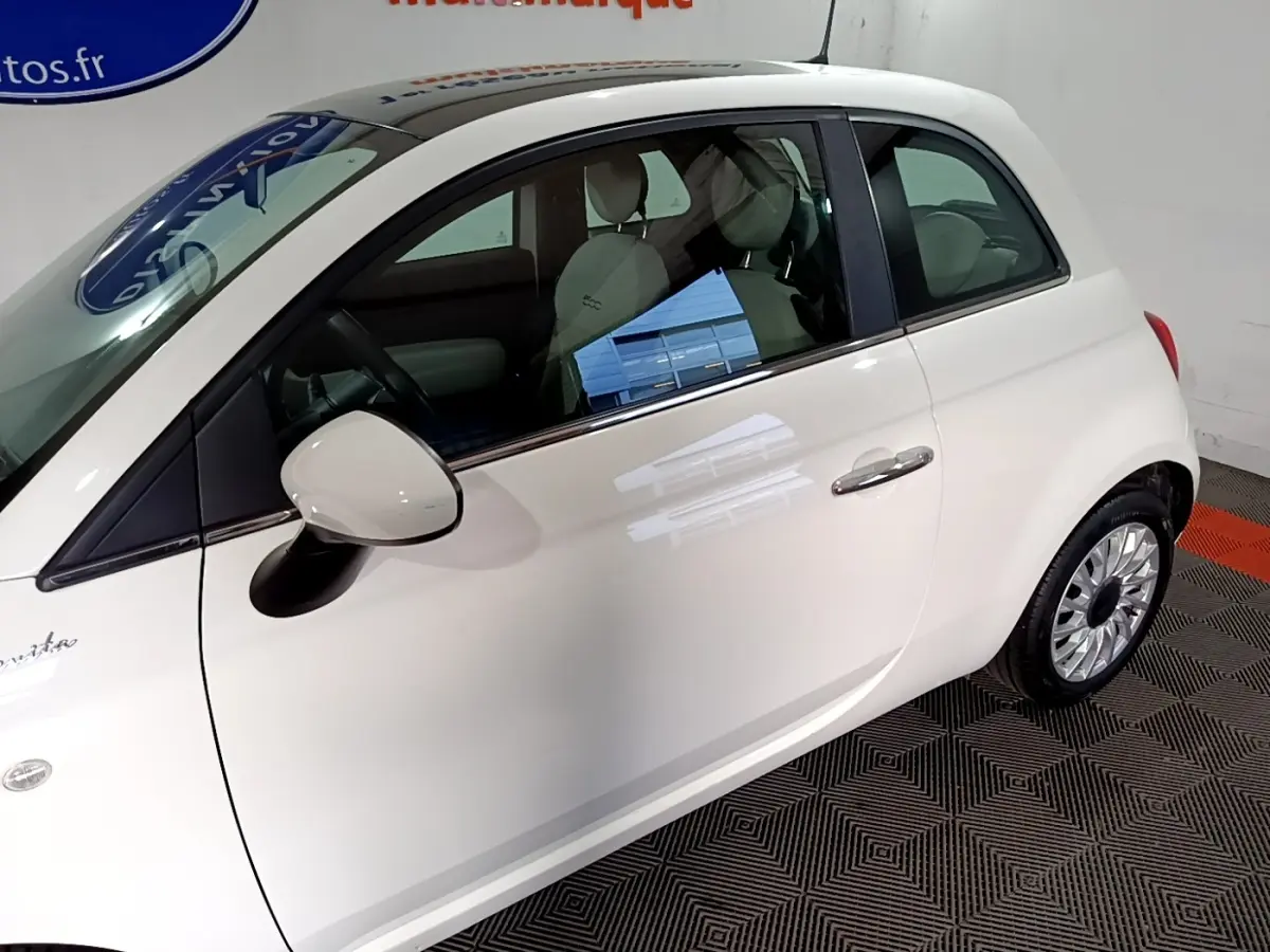 Vue côté gauche d'une Fiat 500 blanche Dolcevita 2021, avec rétroviseur et jante visibles en intérieur showroom.
