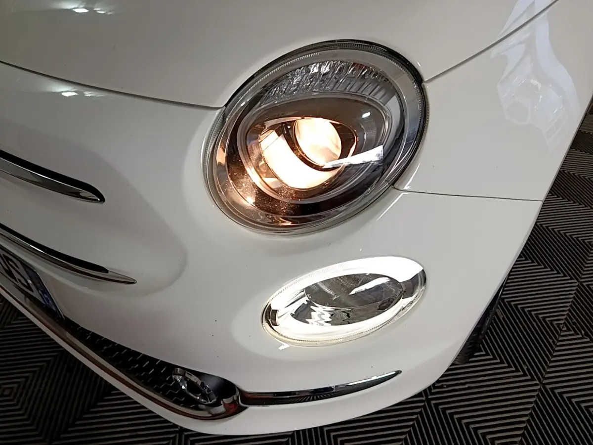Gros plan sur le phare avant droit allumé d'une FIAT 500 blanche, mettant en valeur les détails chromés du pare-chocs.