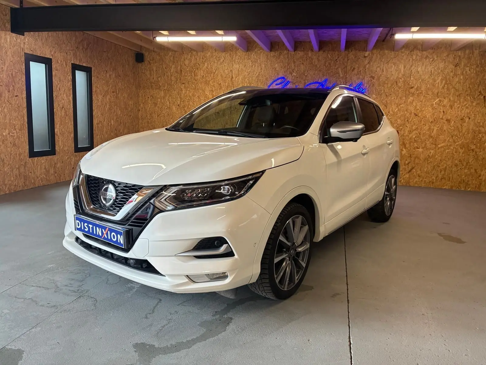Nissan Qashqai blanc en 3/4 avant droit, intérieur visible noir, dans un showroom avec mur en bois et néon bleu.