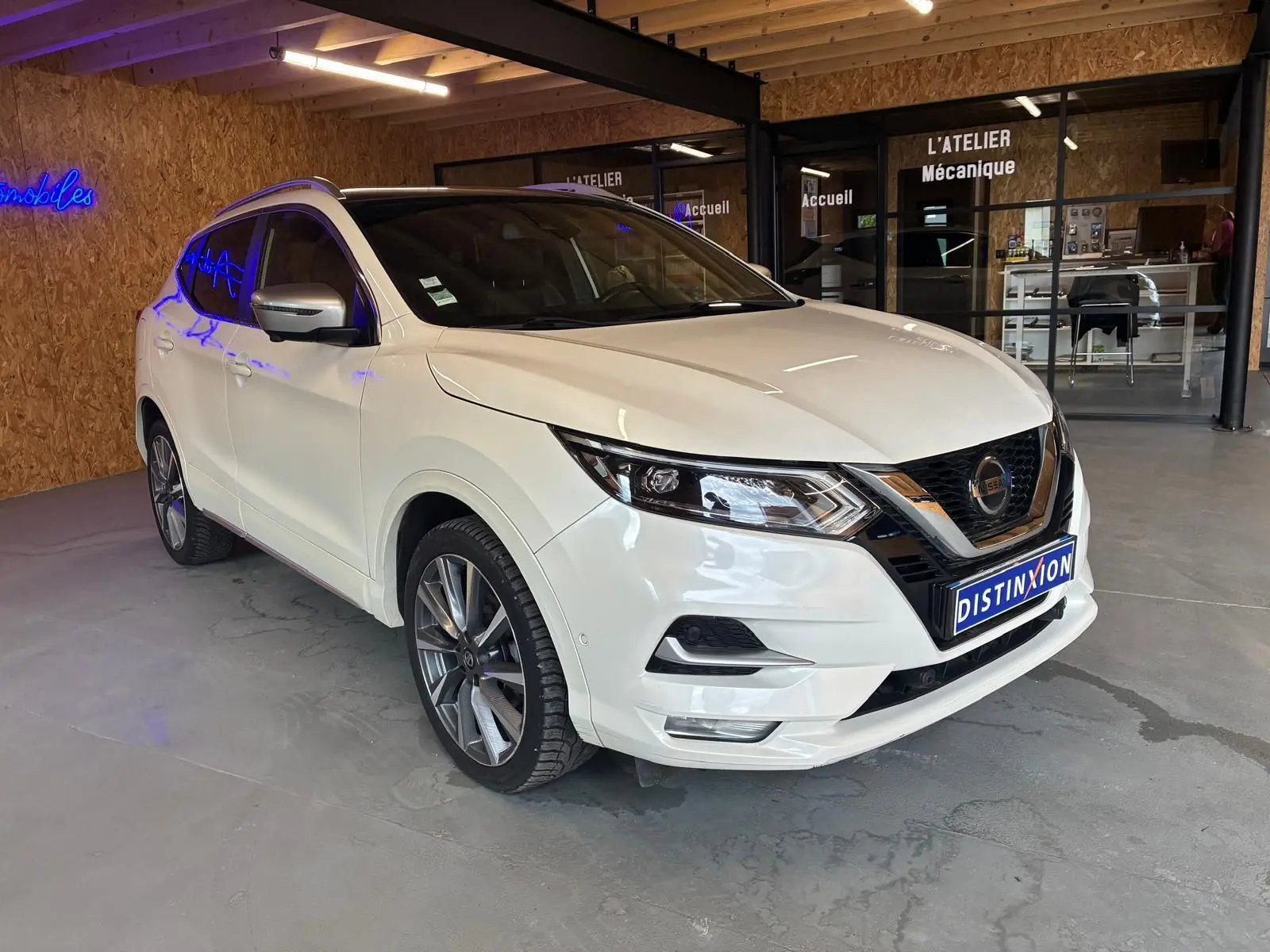 Nissan Qashqai blanc en 3/4 avant droit dans un garage, avec jantes alliage et toit panoramique noir.
