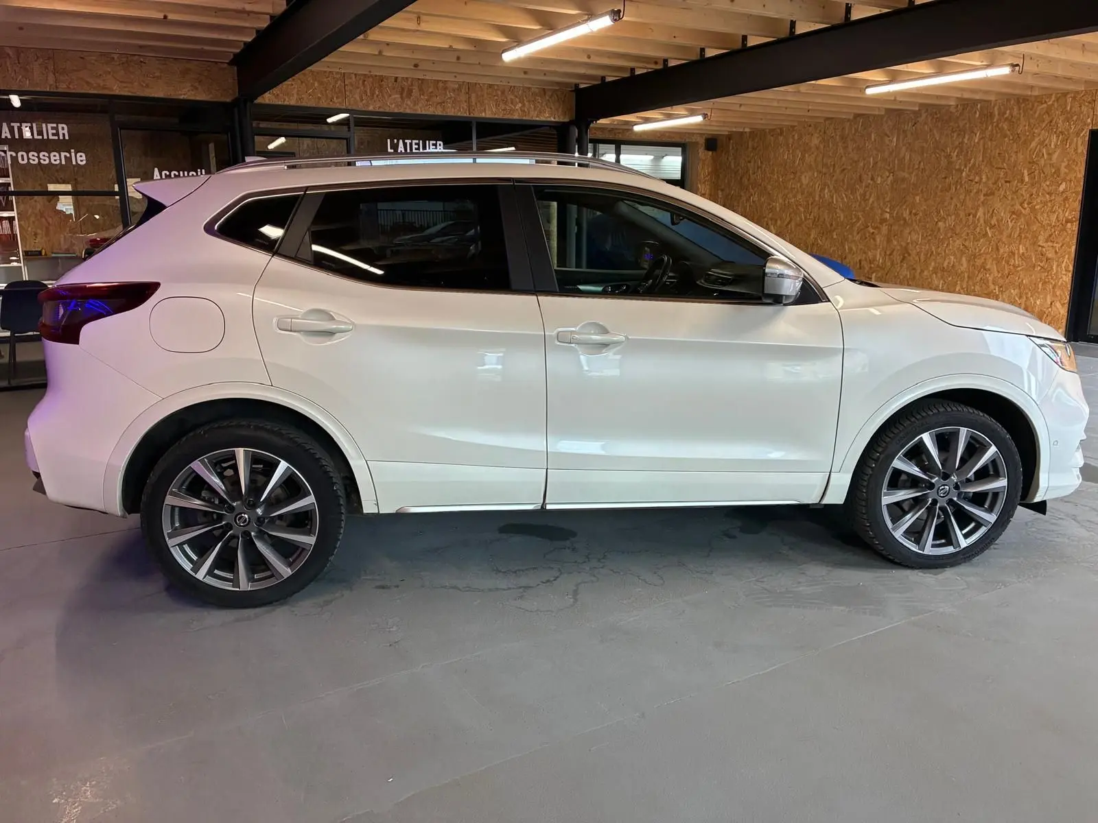 Vue de profil côté gauche d'un Nissan Qashqai blanc 2021 dans un garage, avec jantes alliage et vitres teintées.
