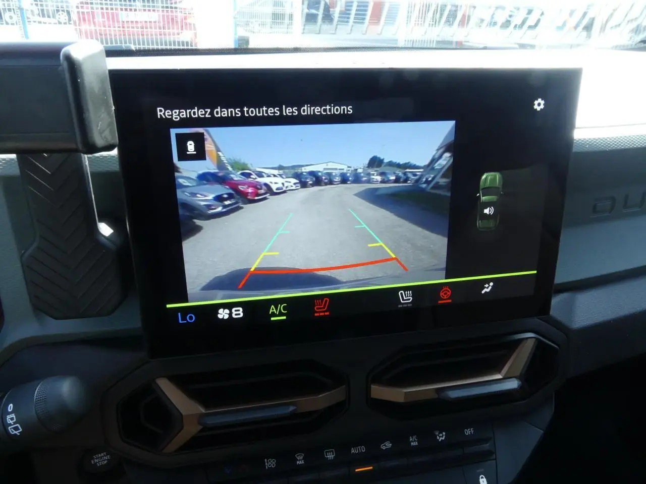 Écran tactile intérieur du Dacia Duster blanc 2025 montrant la caméra de recul avec vue arrière sur un parking.