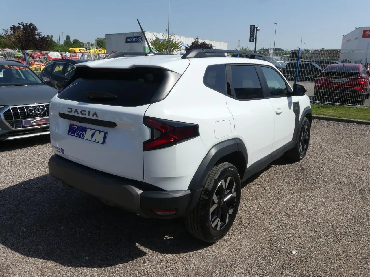 Vue 3/4 arrière droite d'un Dacia Duster blanc 2025 avec jantes alliage noires et barres de toit noires.