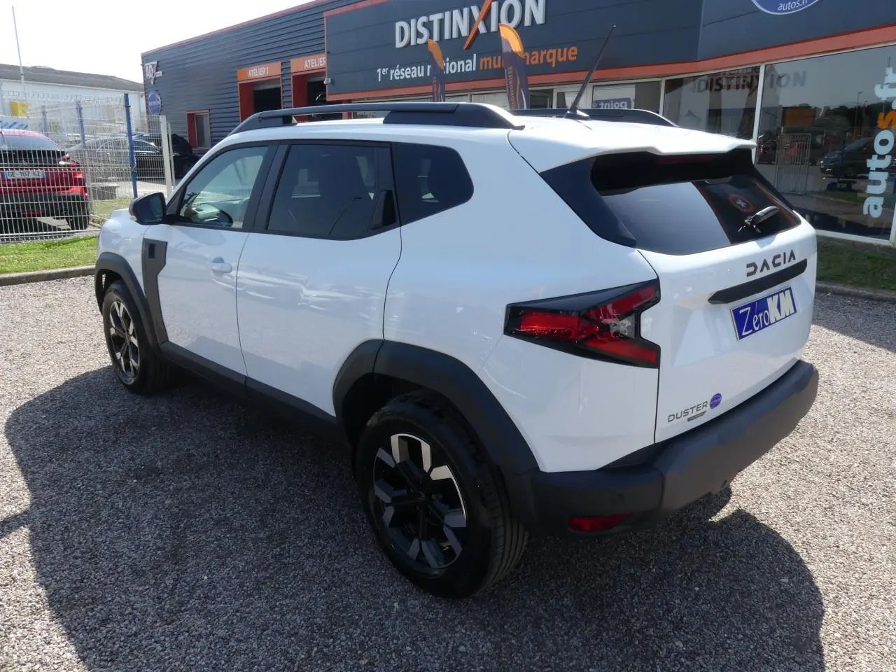 Vue 3/4 arrière droite d'un Dacia Duster blanc 2025 avec jantes alliage noires et barres de toit noires.