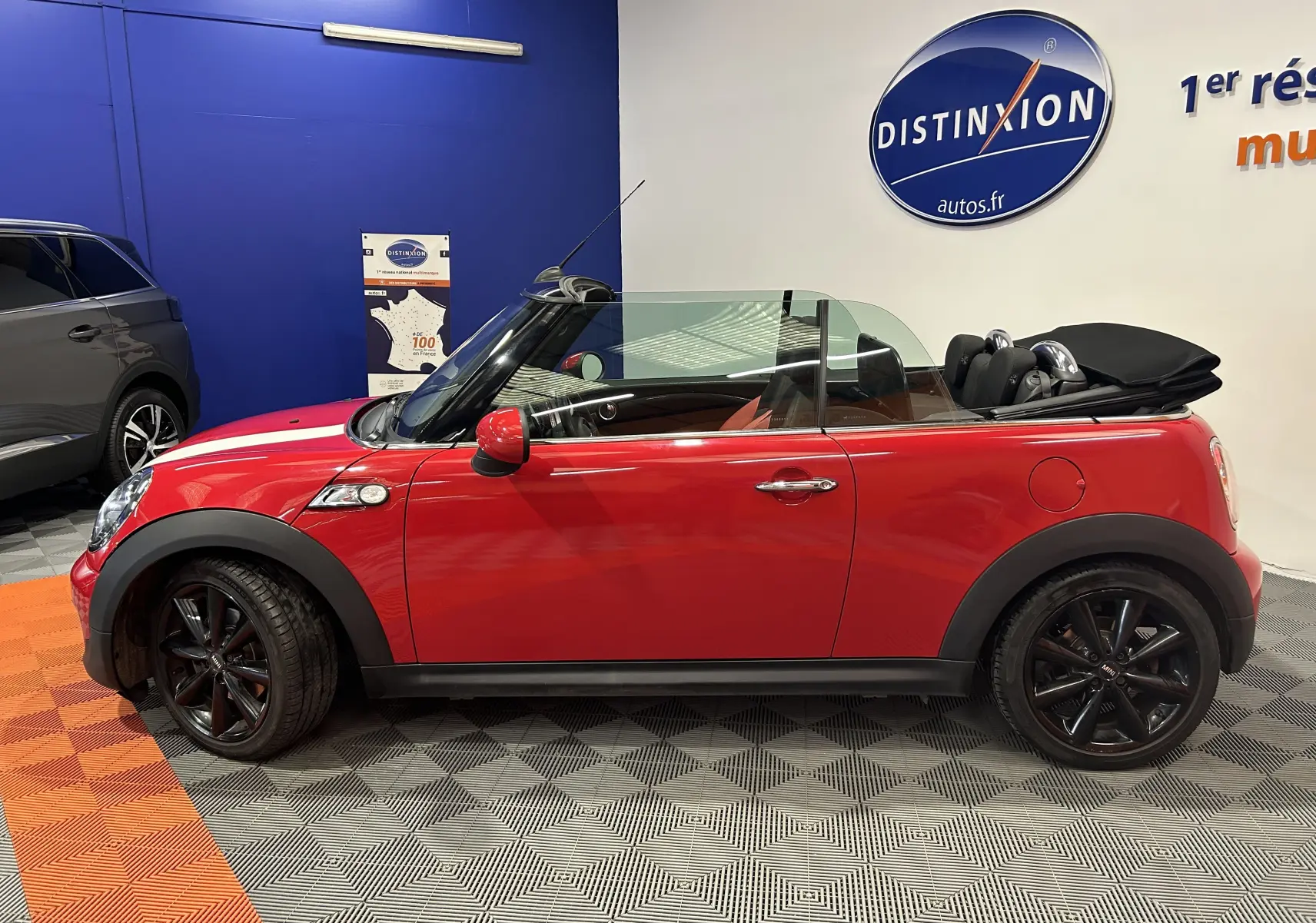 Profil côté gauche d'une MINI Cabriolet Cooper S rouge de 2012, capote noire repliée et jantes noires en intérieur showroom.