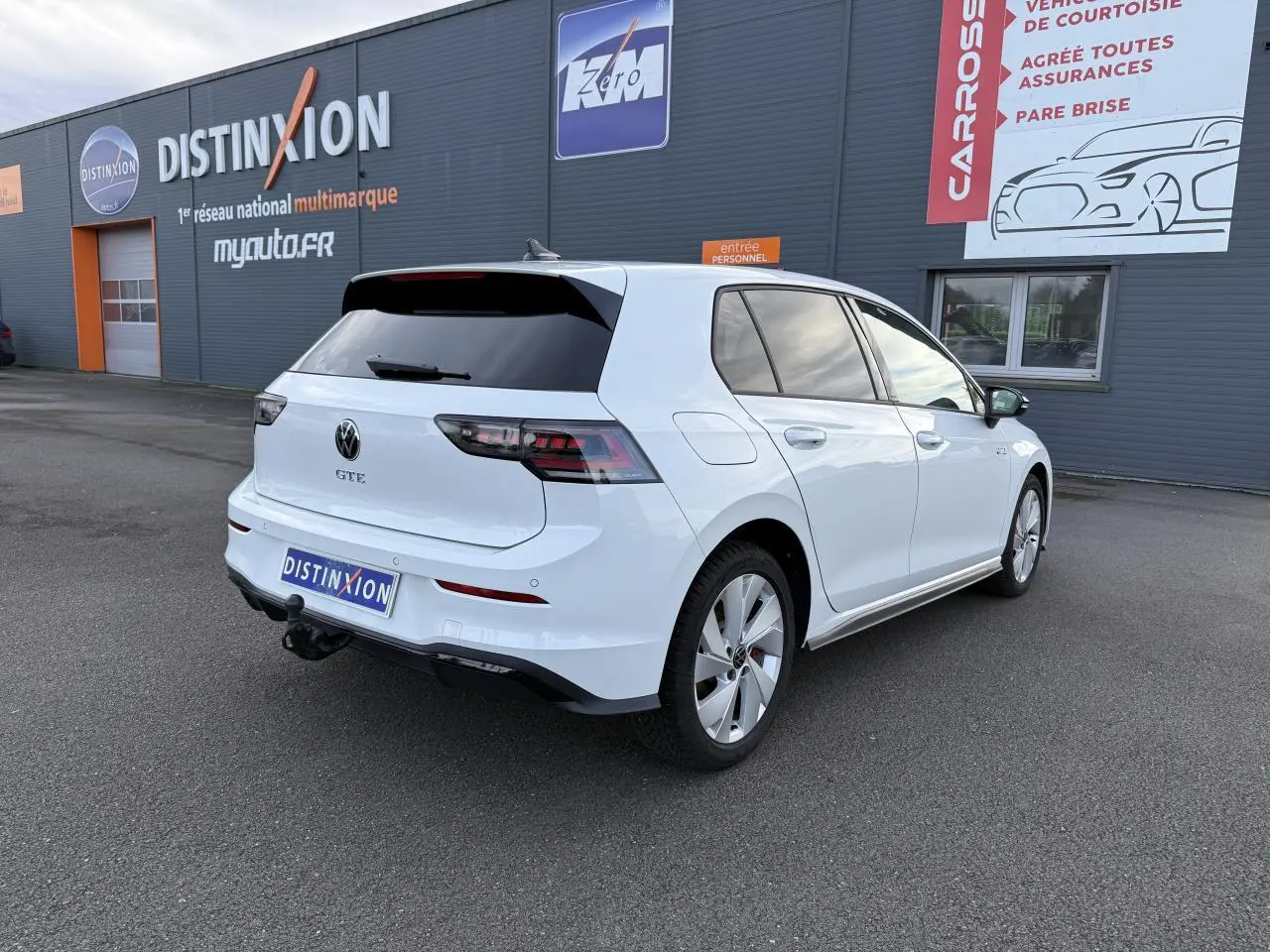 Vue 3/4 arrière droite d'une Volkswagen Golf 1.5 eHybrid blanche nacrée avec attelage et vitres arrière surteintées.