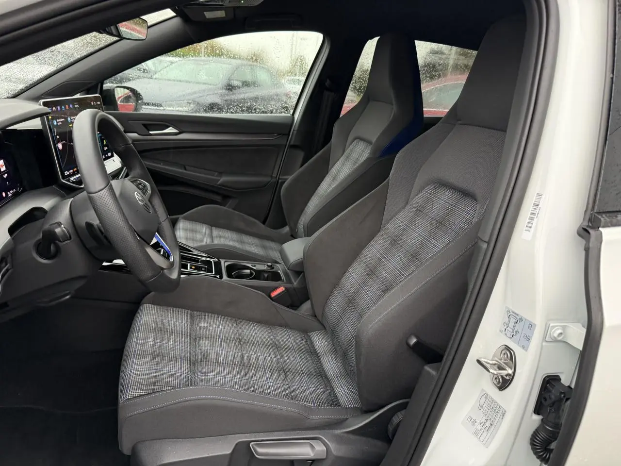 Vue intérieure côté conducteur de la Volkswagen Golf 1.5 eHybrid 2025 avec sièges sport à motif écossais et volant multifonction.