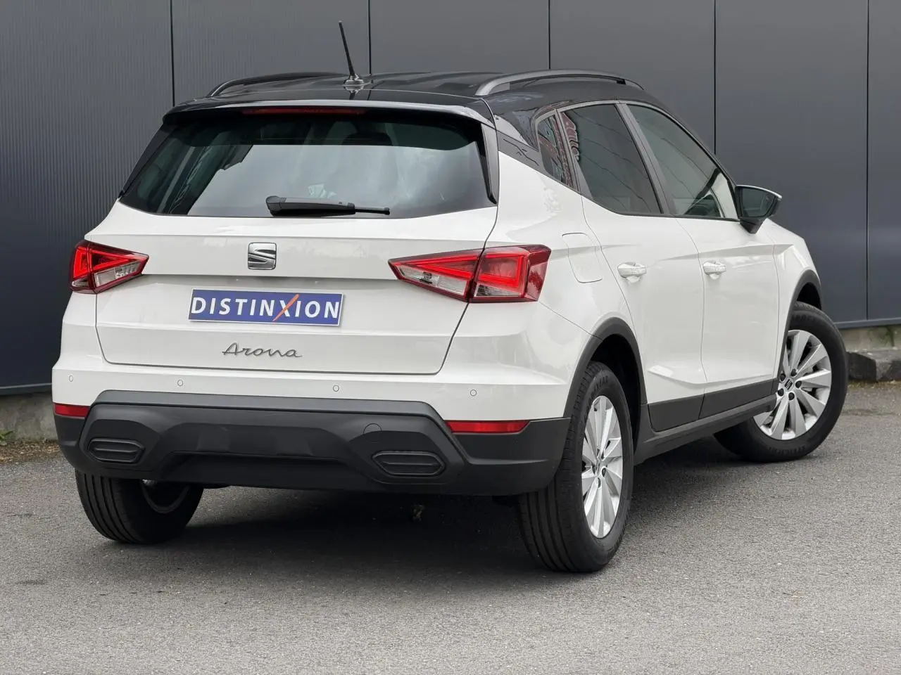 Vue 3/4 arrière droite d'un SEAT Arona blanc avec toit noir et jantes alliage 16 pouces, garé sur sol gris.