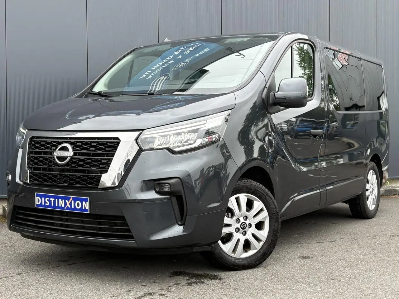 Vue 3/4 avant d’un Nissan Primastar gris comète avec hayon vitré et jantes alliage, stationné devant un mur gris.