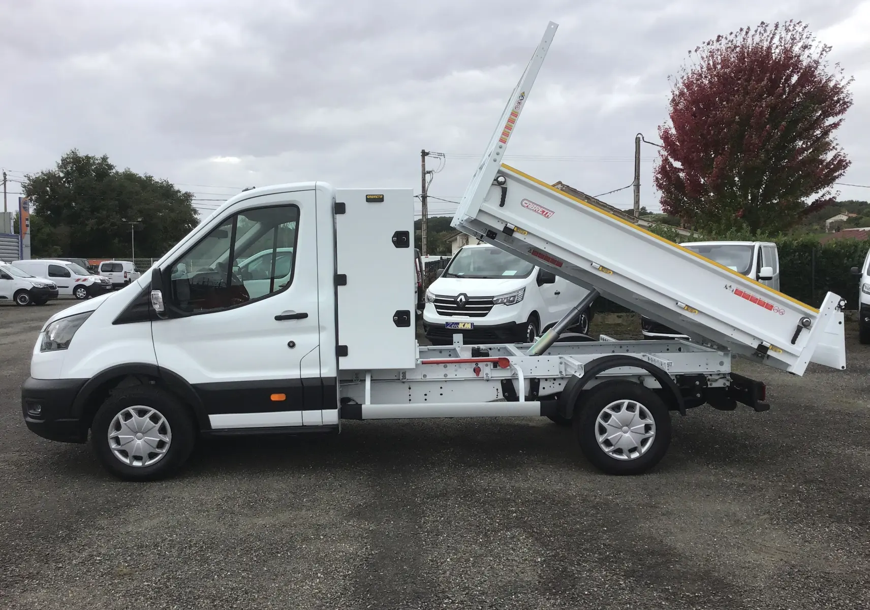 Vue de profil côté gauche d'un Ford Transit blanc avec benne Cabreta relevée et coffre latéral acier.