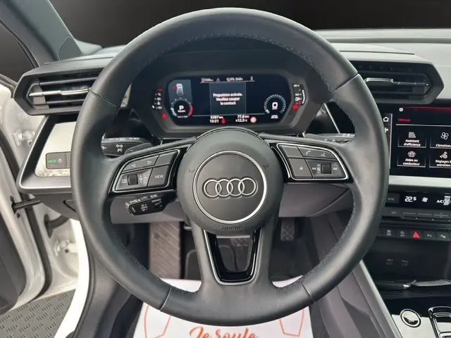 Vue centrée sur le volant cuir noir d'une Audi A3 Sportback blanc Arkona, avec combiné d'instruments digital visible.