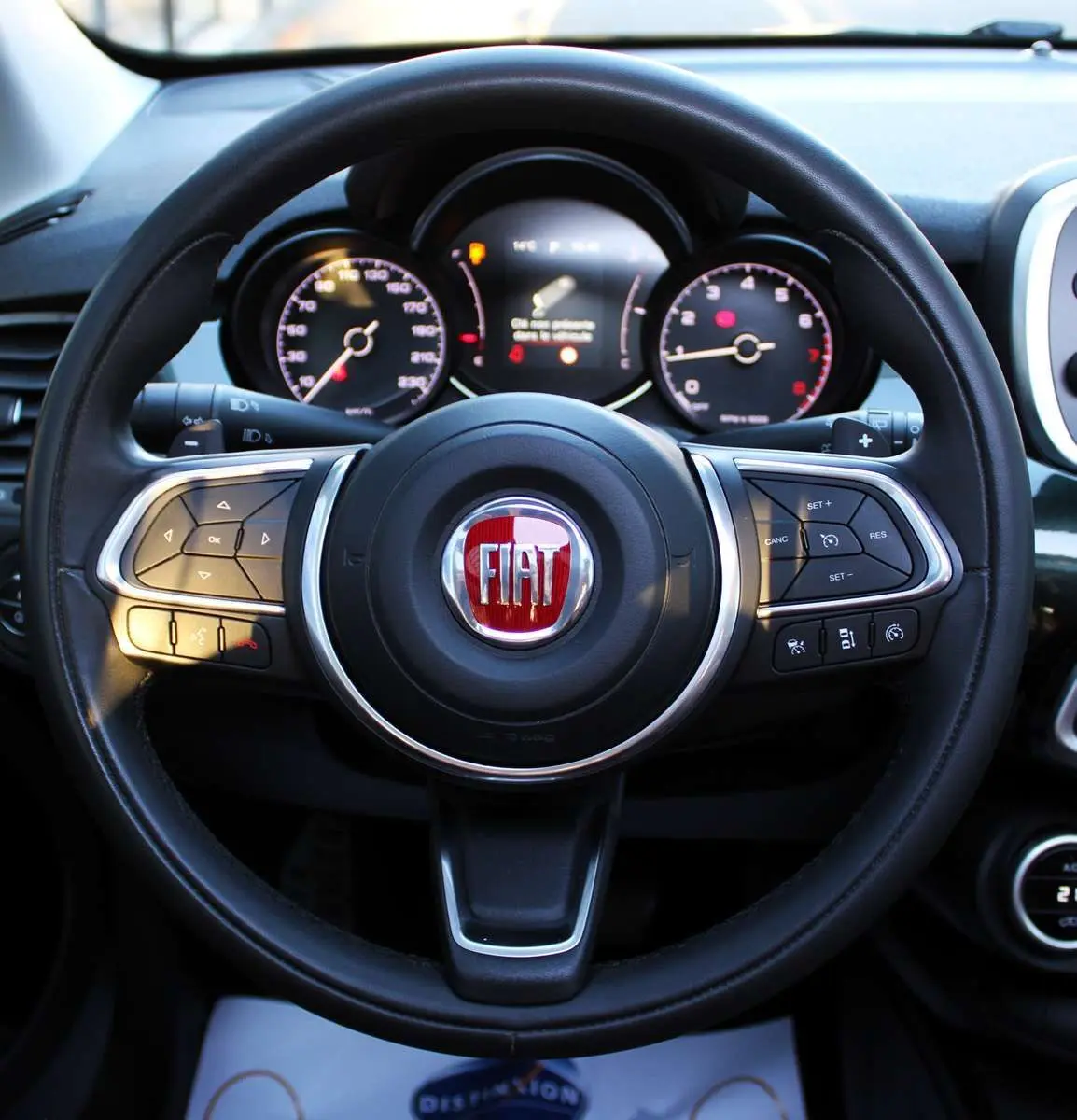 Vue rapprochée du volant noir de la Fiat 500X avec logo rouge et tableau de bord numérique en arrière-plan.