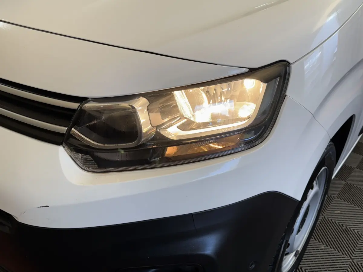 Gros plan sur le phare avant droit allumé d’un Citroën Berlingo blanc avec pare-chocs noir.
