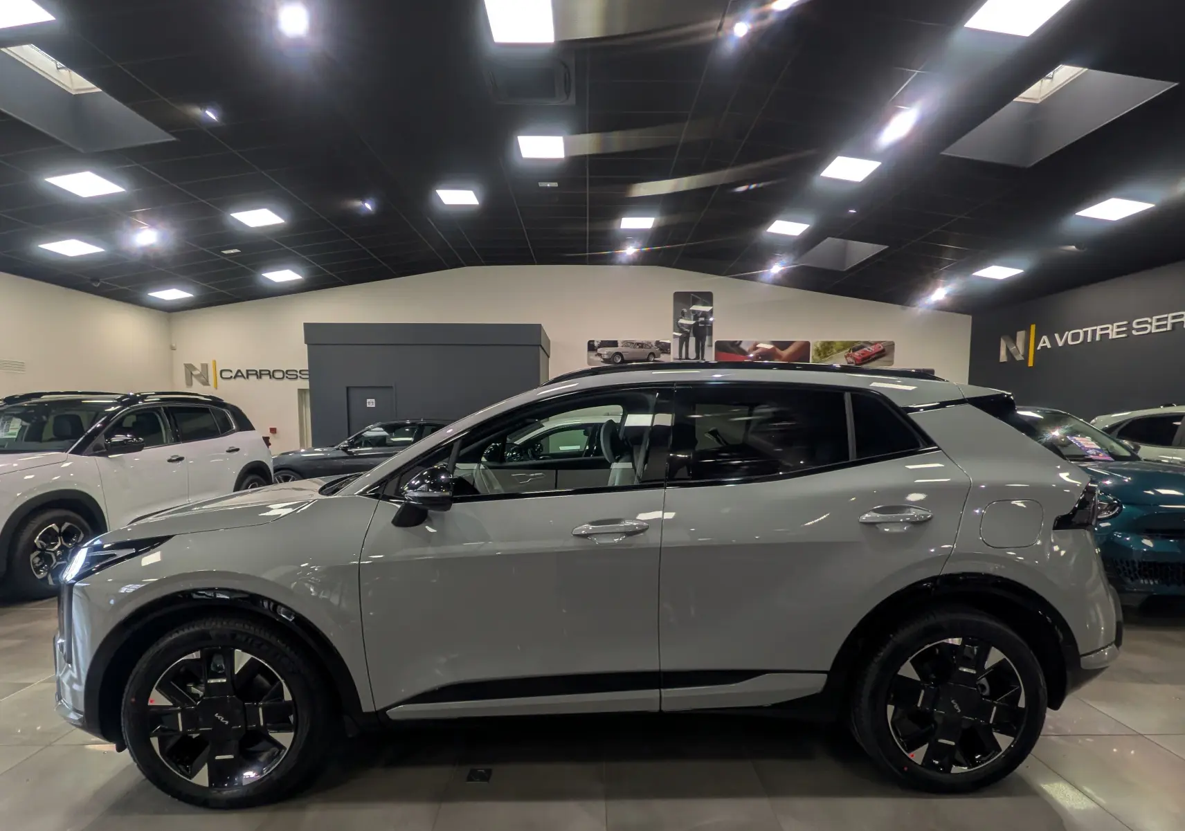 Vue de profil côté gauche d'un KIA SPORTAGE gris céramique avec toit noir, roues noires et intérieur visible en showroom.
