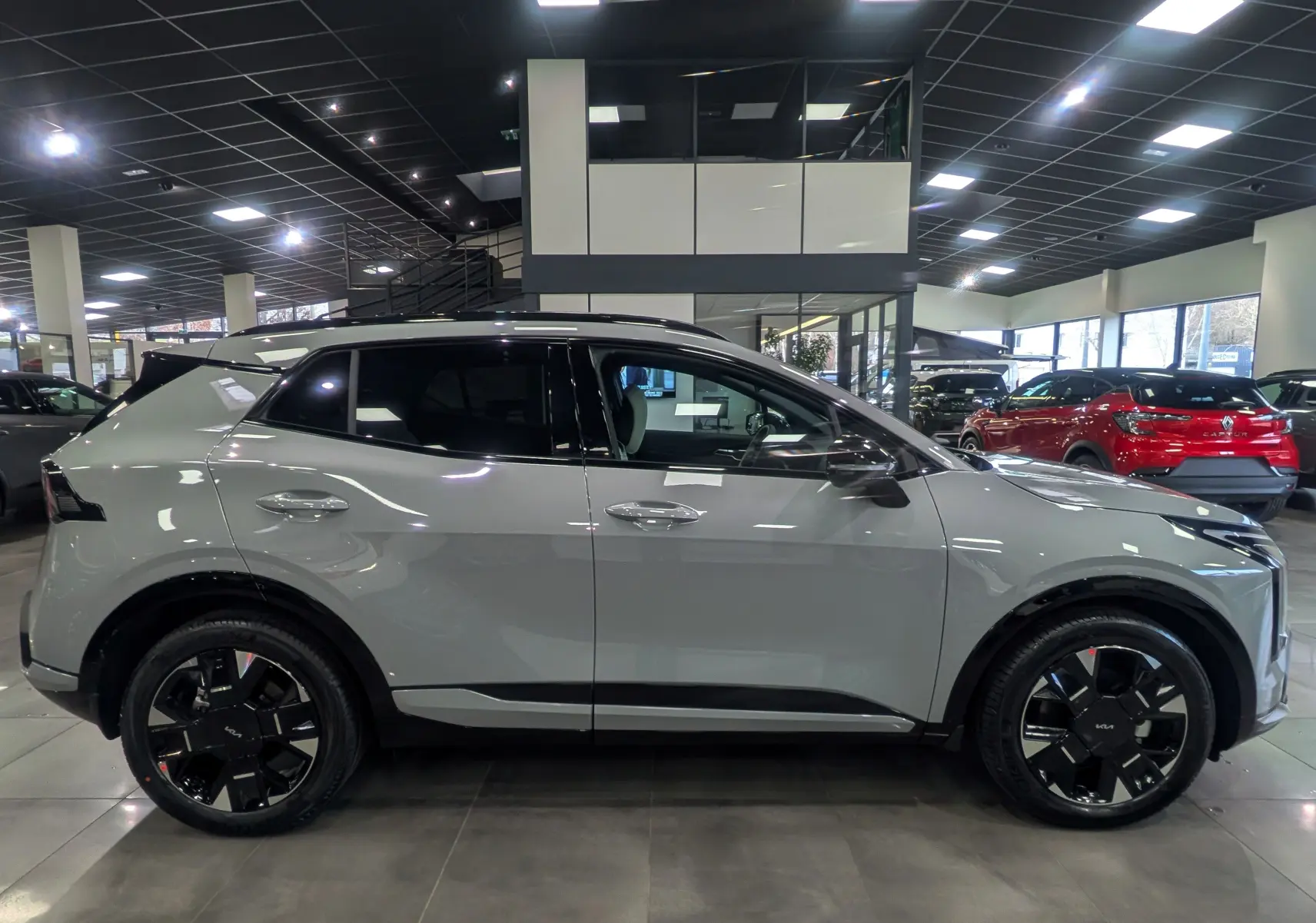 KIA SPORTAGE 2025 gris céramique avec toit noir, vue de profil côté gauche, jantes noires distinctives en showroom.
