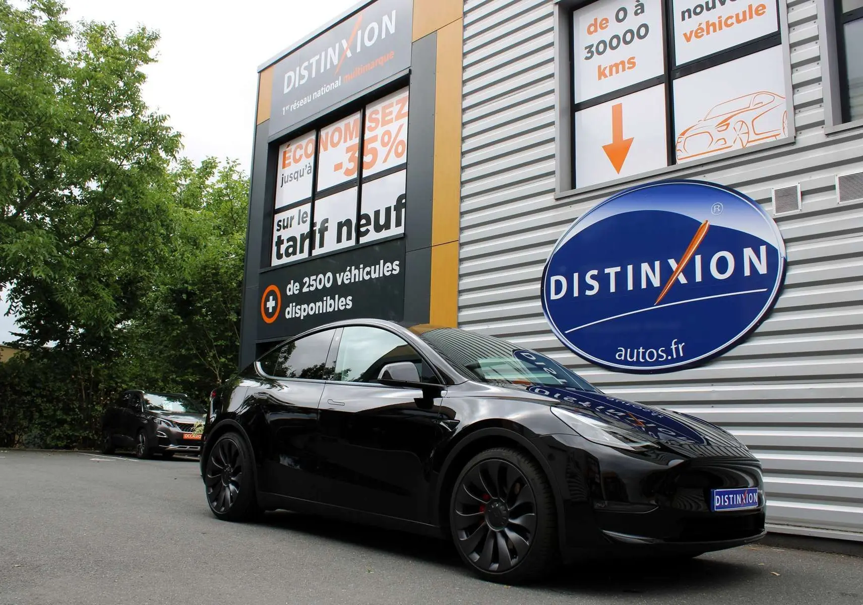 Tesla Model Y noir métal en 3/4 avant droit, garée devant un mur avec logo Distinxion bleu et orange.