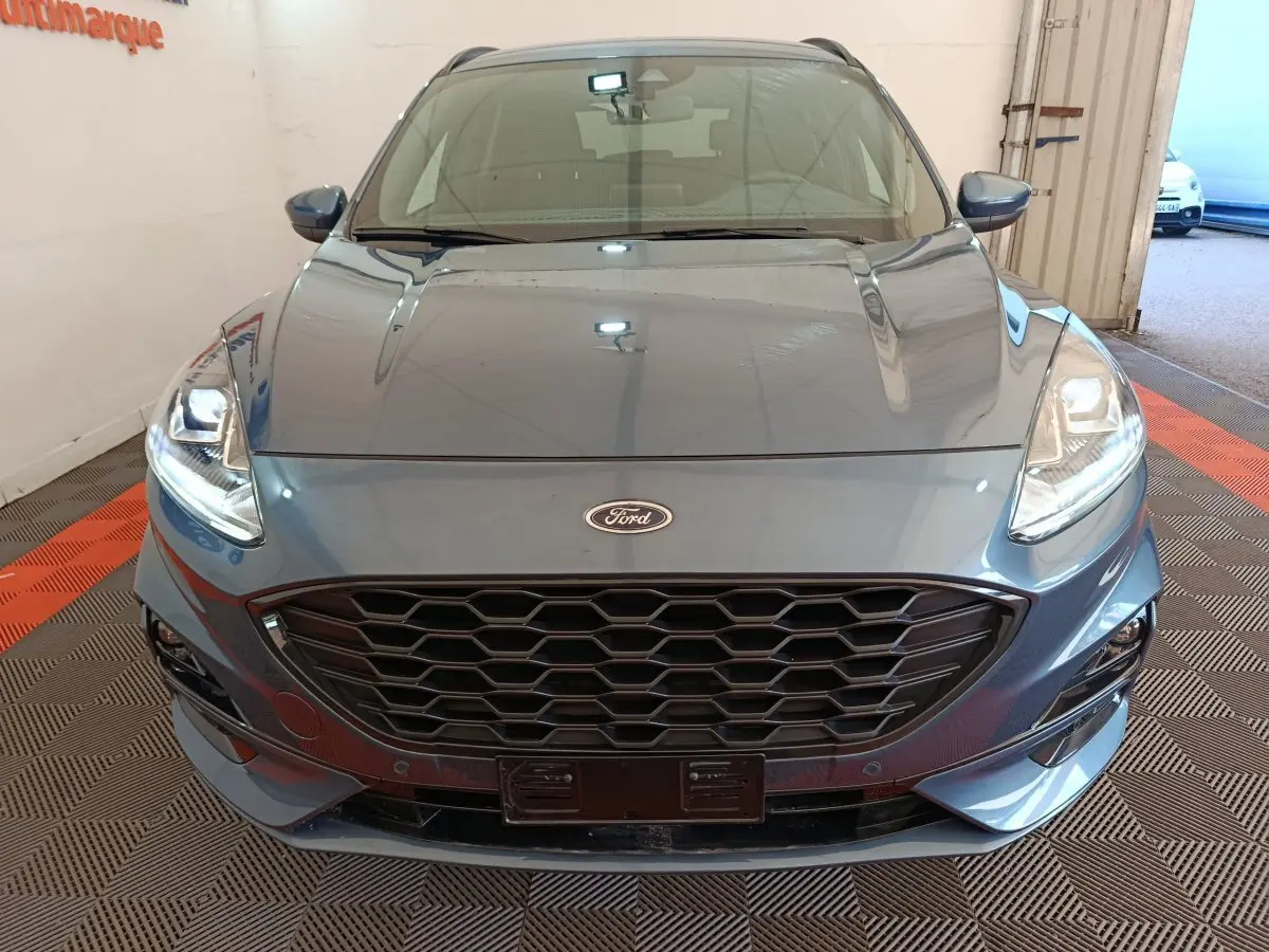 Vue frontale rapprochée du Ford Kuga bleu 2024 avec calandre noire et phares allumés en intérieur.