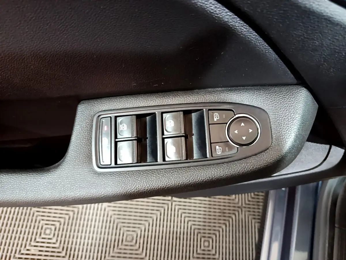 Gros plan sur les commandes des vitres électriques et rétroviseurs sur la porte côté conducteur d’une Renault Clio gris foncé.