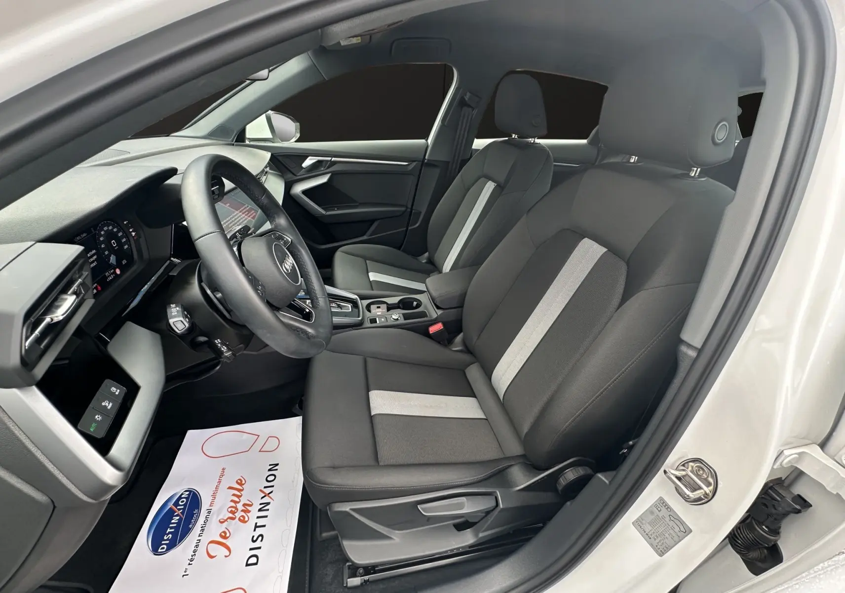 Intérieur côté conducteur d'une Audi A3 Sportback blanche, sièges tissu noir avec bande grise centrale et volant cuir multifonctions.