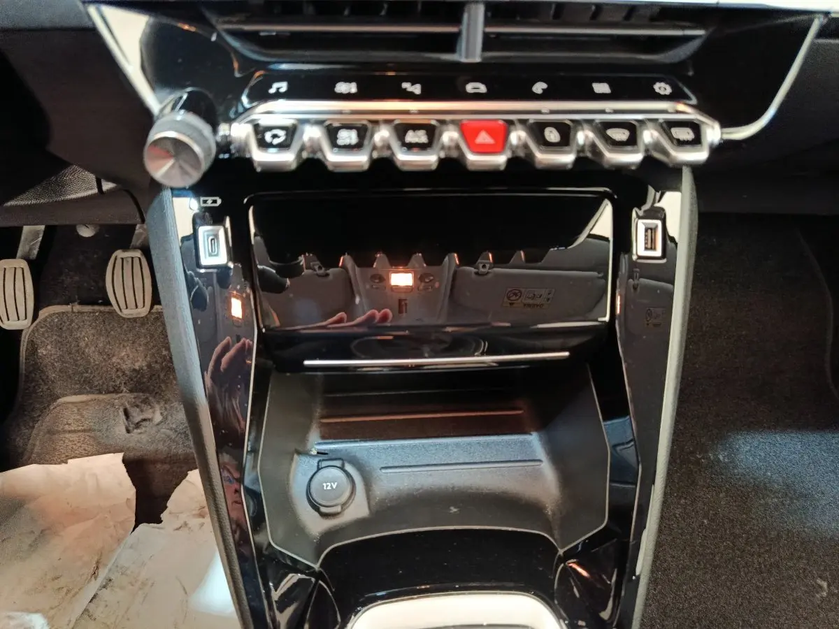 Vue rapprochée de la console centrale noire brillante de la Peugeot 208 BlueHDi 100, avec boutons et prise 12V visible.
