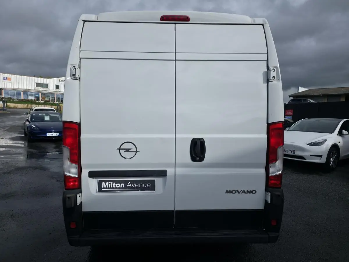 Vue arrière d'un fourgon blanc Opel Movano avec portes battantes et logo visible sous un ciel nuageux.