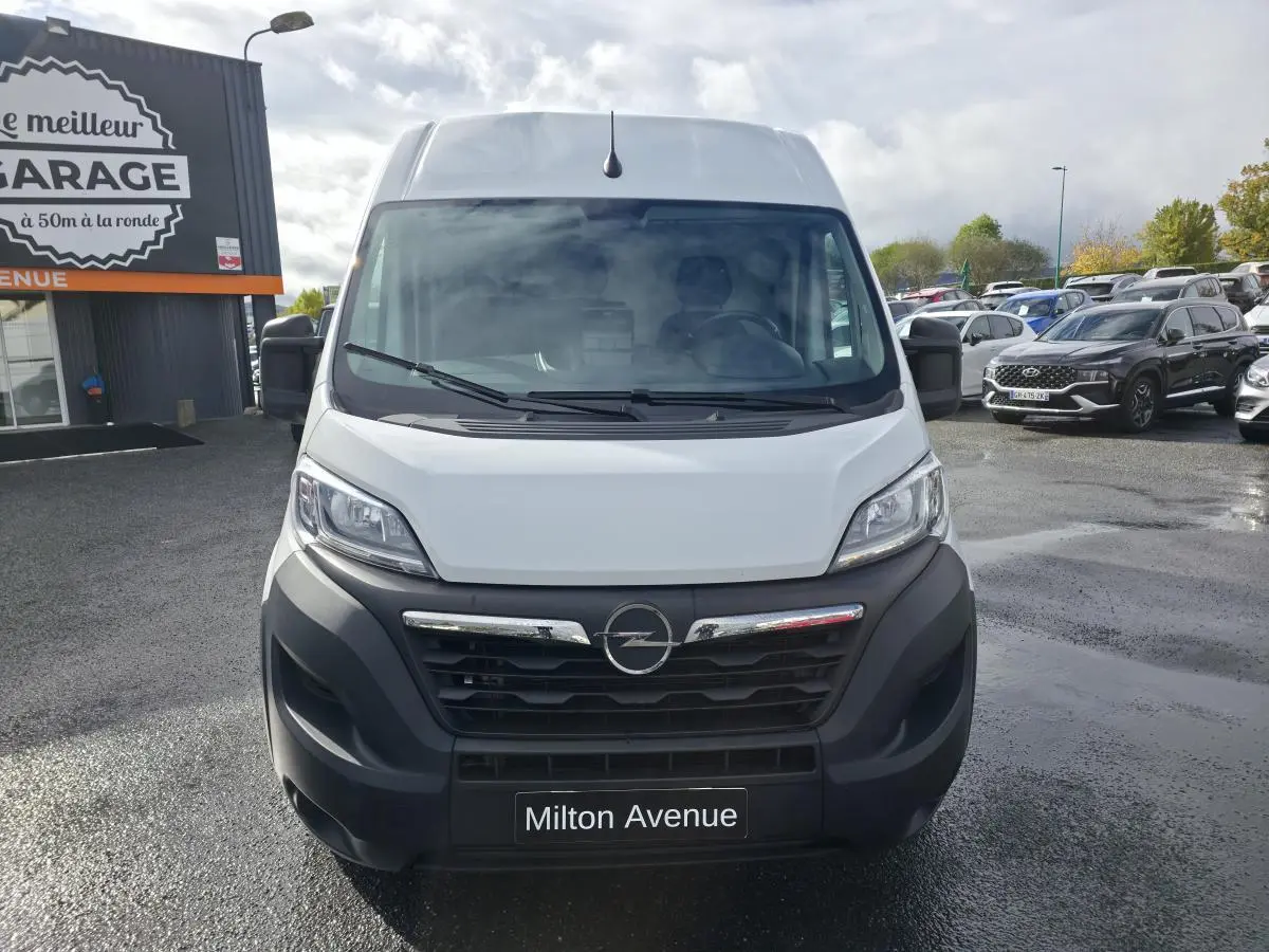 Vue de face d'un fourgon Opel Movano blanc avec calandre noire et logo Opel au centre, stationné devant un garage.