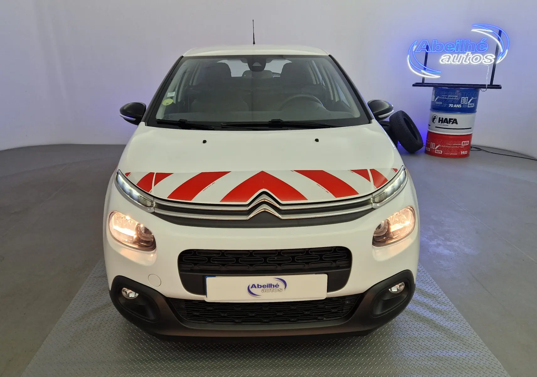 Vue avant d'une Citroën C3 Société blanche avec bandes rouges diagonales sur le capot et phares allumés en intérieur.
