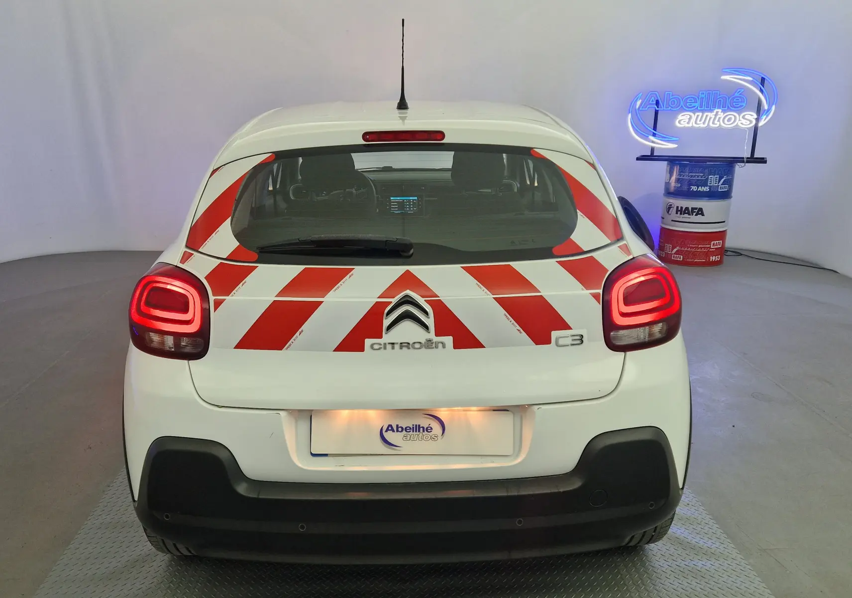 Vue arrière d'une Citroën C3 blanche 2019 avec bandes rouges diagonales de signalisation et feux arrière allumés.