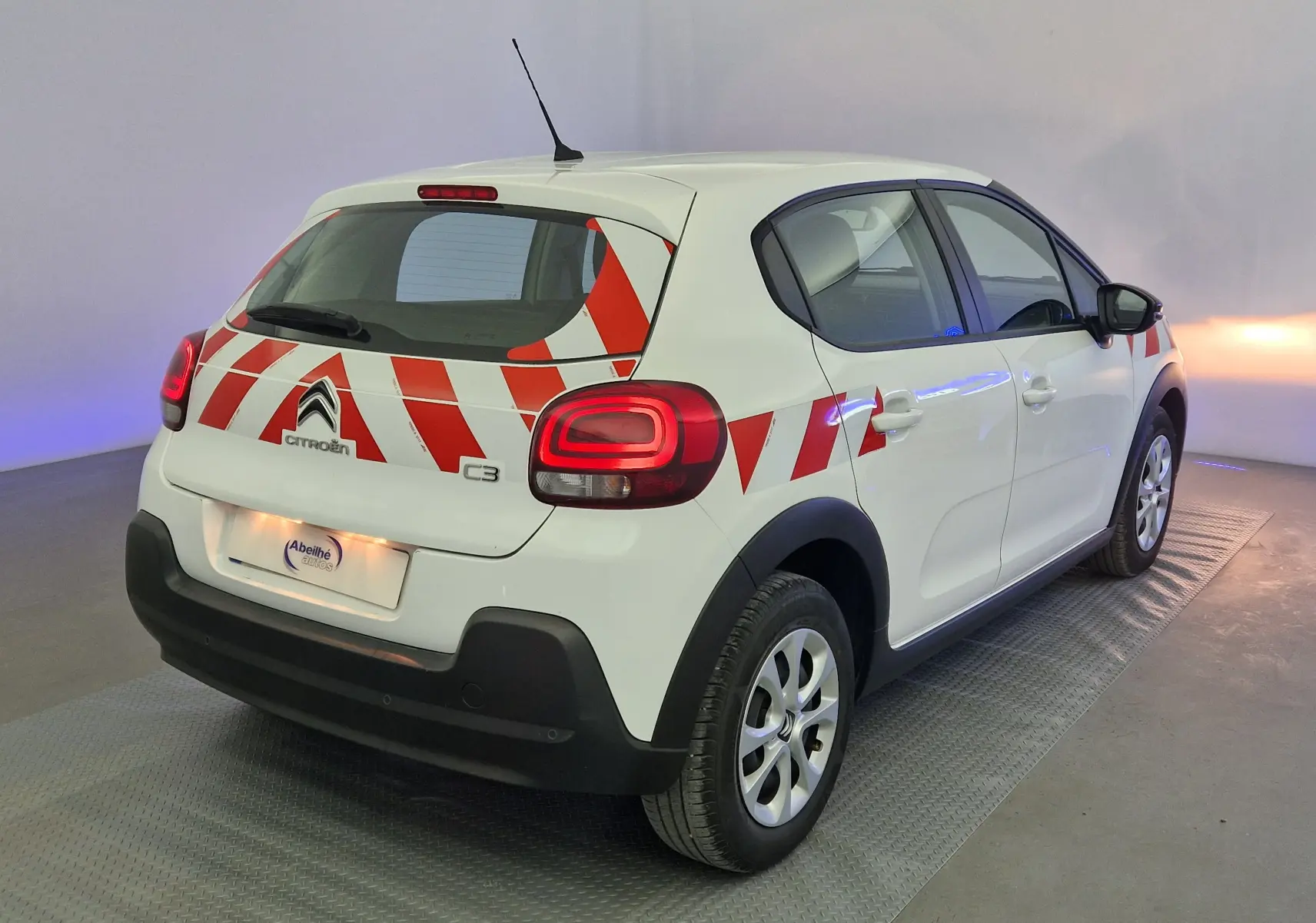 Vue 3/4 arrière droite d'une Citroën C3 blanche avec bandes rouges diagonales distinctives sur la carrosserie.