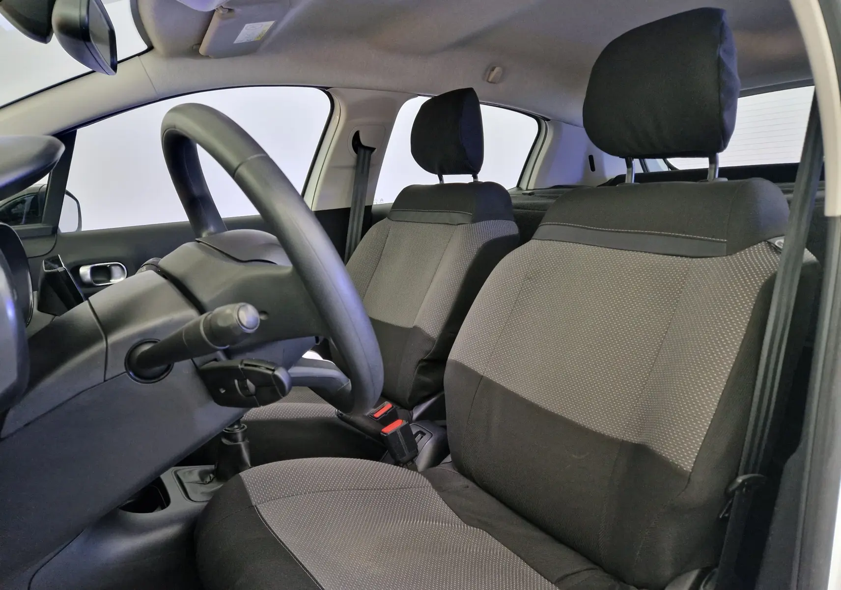 Vue intérieure côté conducteur de la Citroën C3 Société 2019, sièges tissu gris et noir avec volant et levier de vitesse visibles.