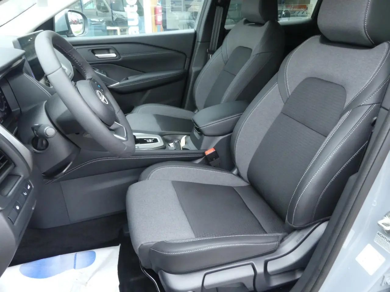 Vue intérieure côté conducteur du Nissan Qashqai 1.5 e-Power 2025 avec sièges gris en tissu et accoudoir central.