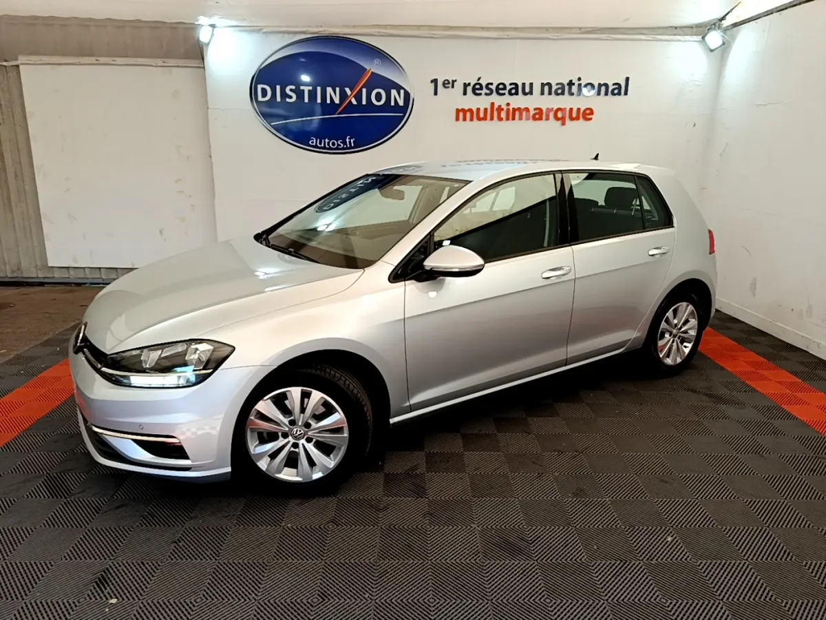 Volkswagen Golf 1.6 TDI gris clair vue 3/4 avant droit dans un showroom avec logo Distinxion en arrière-plan.