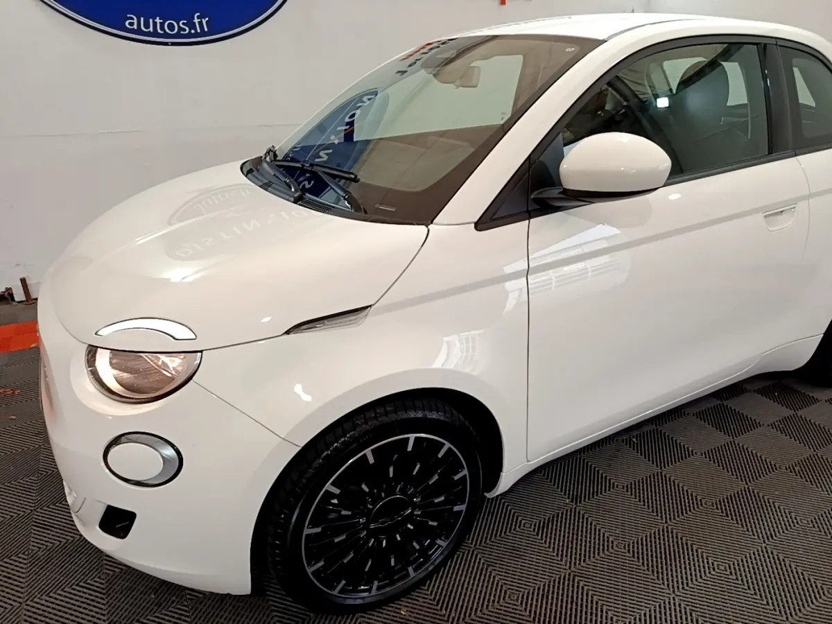 Vue 3/4 avant gauche d'une FIAT 500 électrique blanche avec jantes noires et phares allumés.